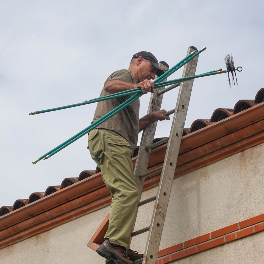 A man climbs the ladder to the roof with a chimney brush in his hand.