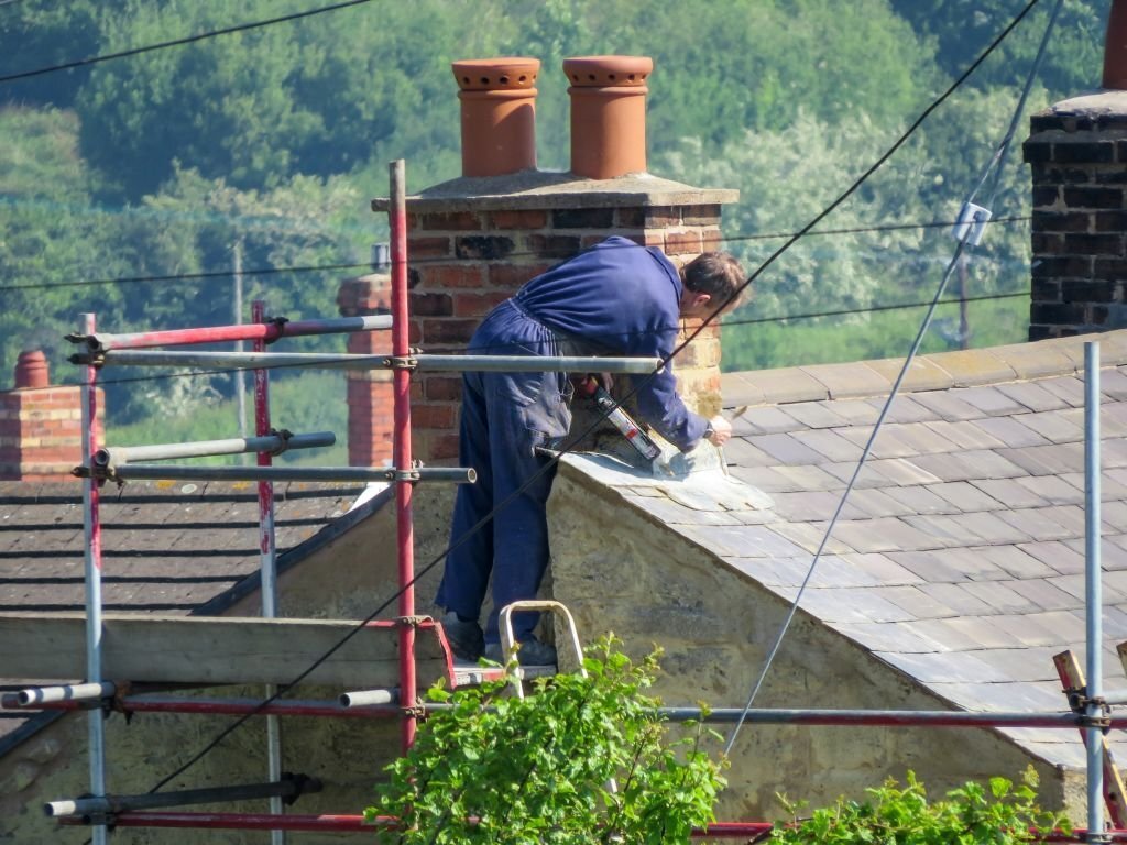 Wrexham UK - May 25, 2017: Roofer repairing flashing on chimney stack on a slate roof of a domestic house. Standing on scaffolding. Close up, high angle point of view. Countryside background.