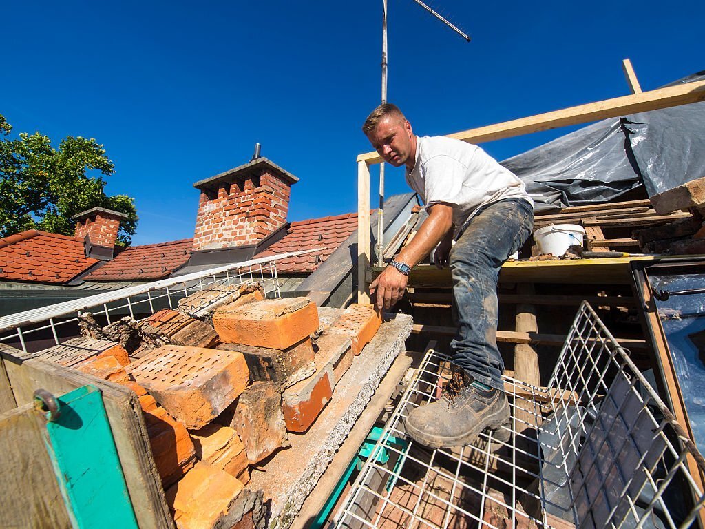 Partial renovation of the roof brick chimneys of an old house in Ljubljana. Man is loading construction lift, with old bricks, while in background is some scaffolding that has been erected around a brick chimney stack and the wooden rafters / beams of roofing,